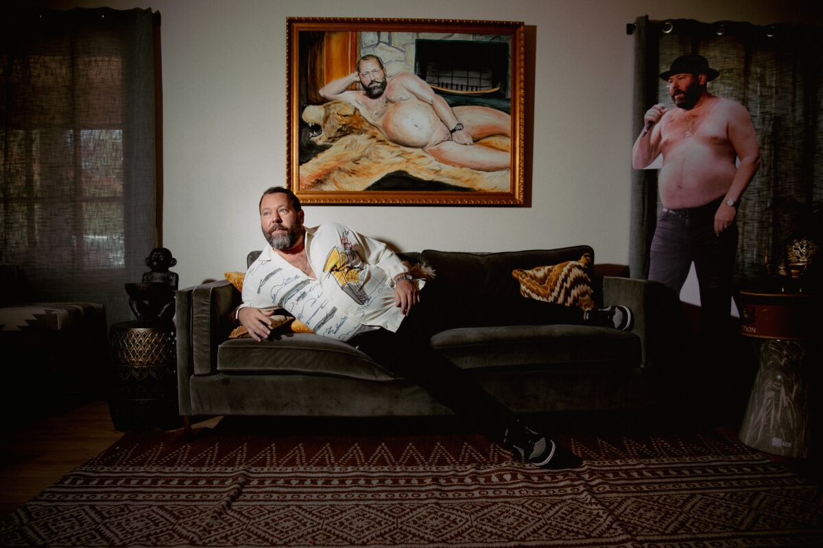 Bert Kreischer on a couch beneath a painting of himself, next to a cardboard cutout of himself.